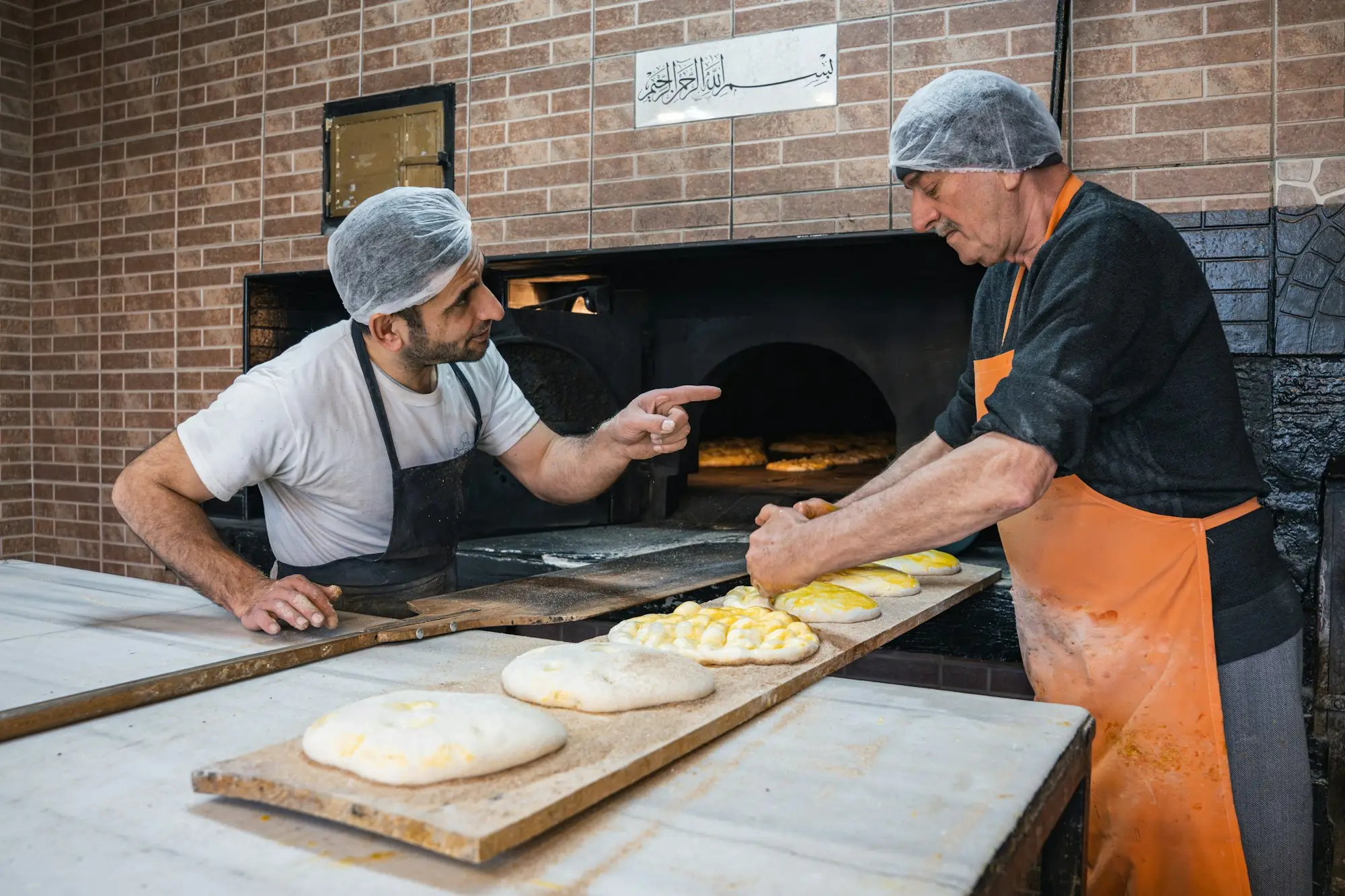 Two bakers preparing dough beside a brick oven in a working bakery